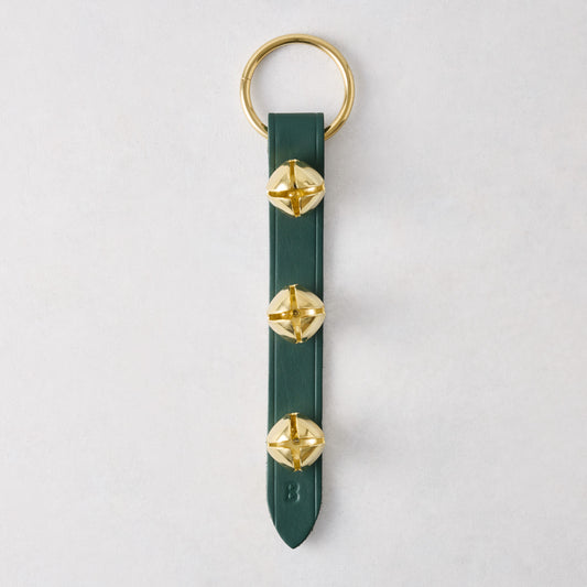 Three gold jingle bells attached to a dark green leather strap with a gold metal ring on a white background.