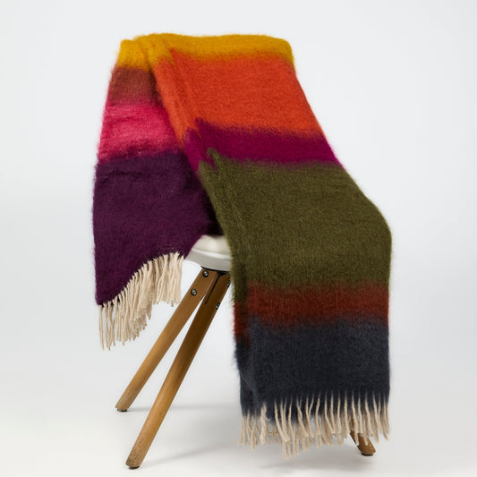 A fuzzy striped wool blanket with cream fringe, draped over a wooden stool against a plain white studio background.