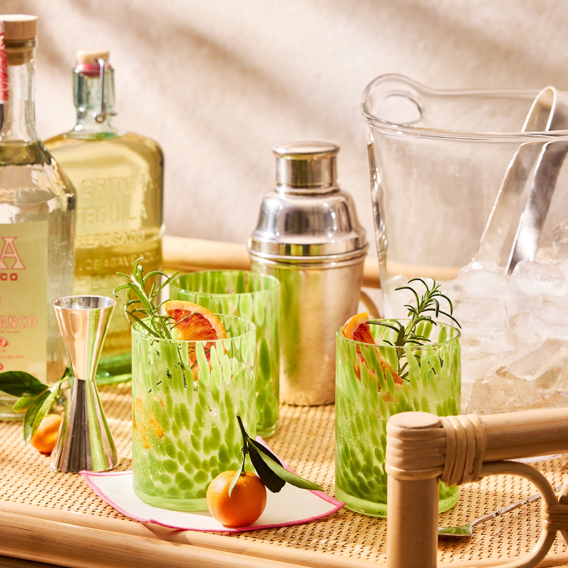 Two green spotted double old fashioned glasses with cocktails, tequila bottles, and bar tools on a rattan tray.