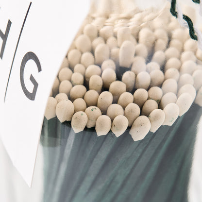 Oversized green matches with white tips in a clear glass bottle, cork topper, and white label.