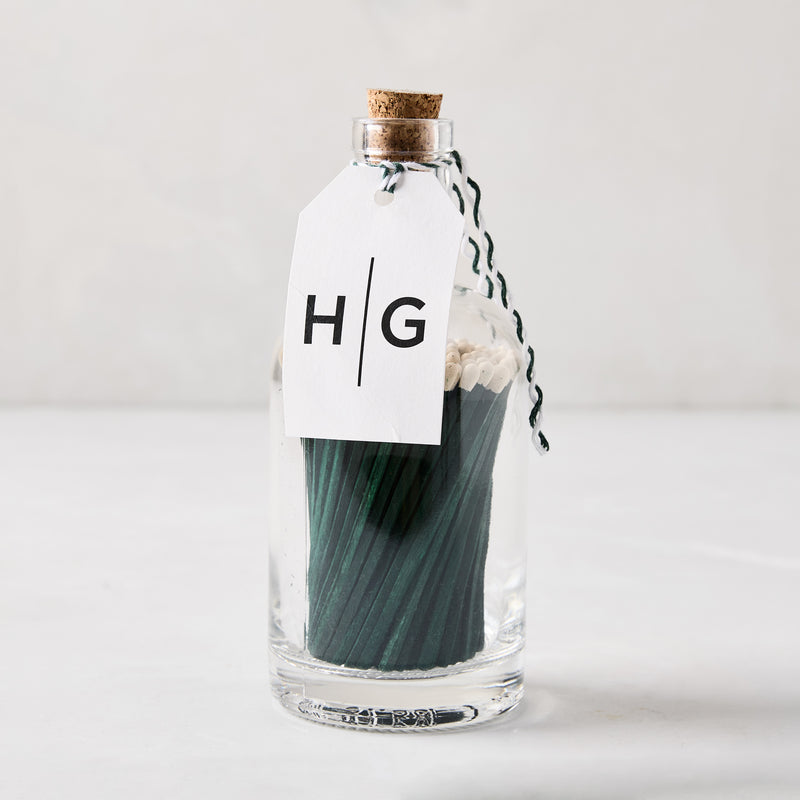 Green with white tip oversized matches in a clear glass bottle with a cork stopper and HG tag.