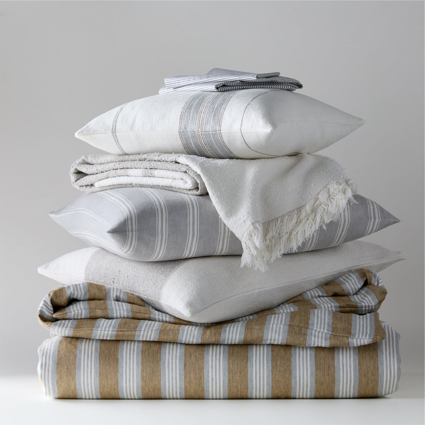 Stack of Guest House Stripe Linen Pillow Shams and bedding in oyster, light grey, and brown stripes.