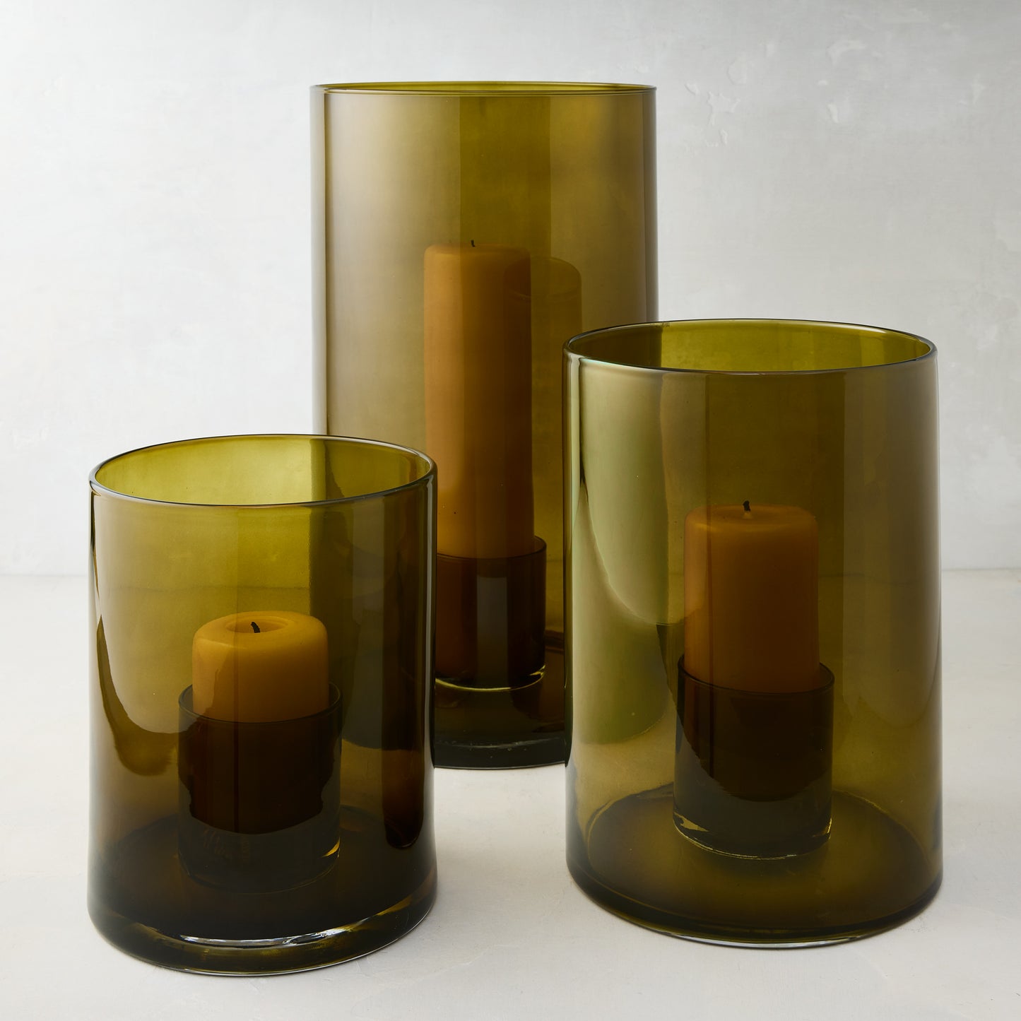 Three handcrafted olive green hand-blown glass hurricanes of varying sizes, each with a pillar candle.