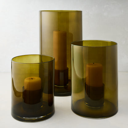 Three handcrafted olive green hand-blown glass hurricanes of varying sizes, each with a pillar candle.