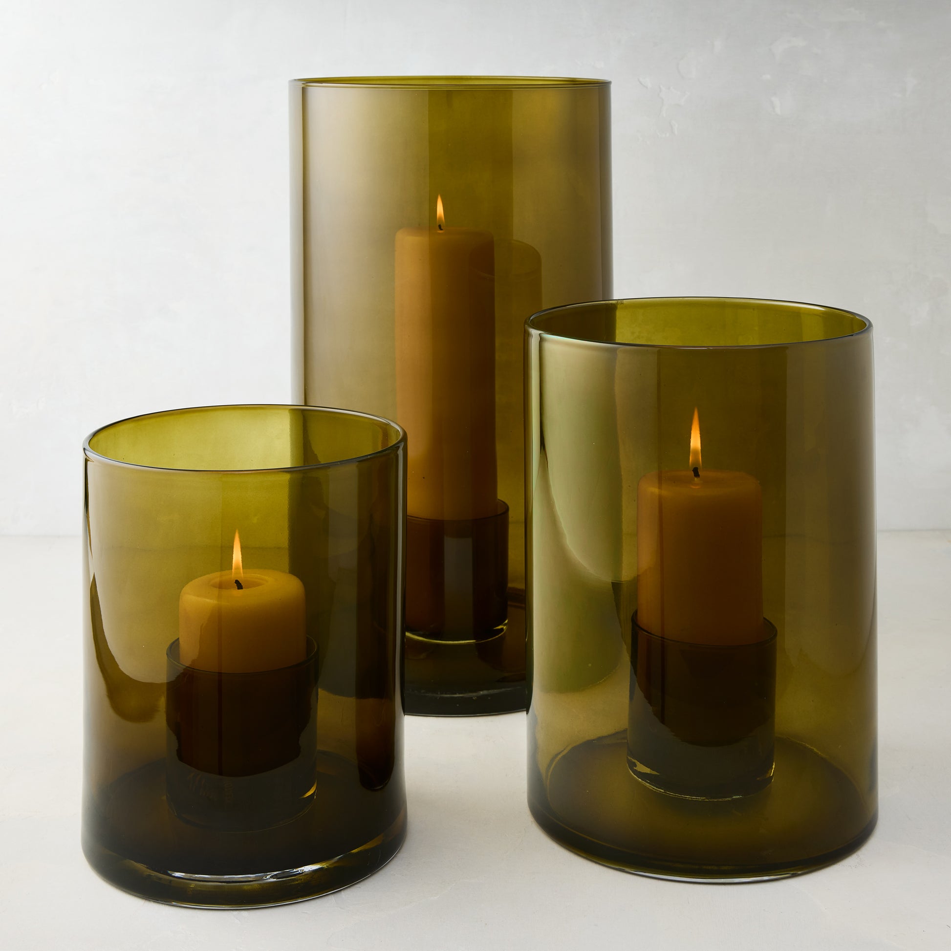Three handcrafted olive green hand-blown glass hurricanes of varying sizes, each with a lit pillar candle.