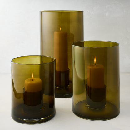 Three handcrafted olive green hand-blown glass hurricanes of varying sizes, each with a lit pillar candle.