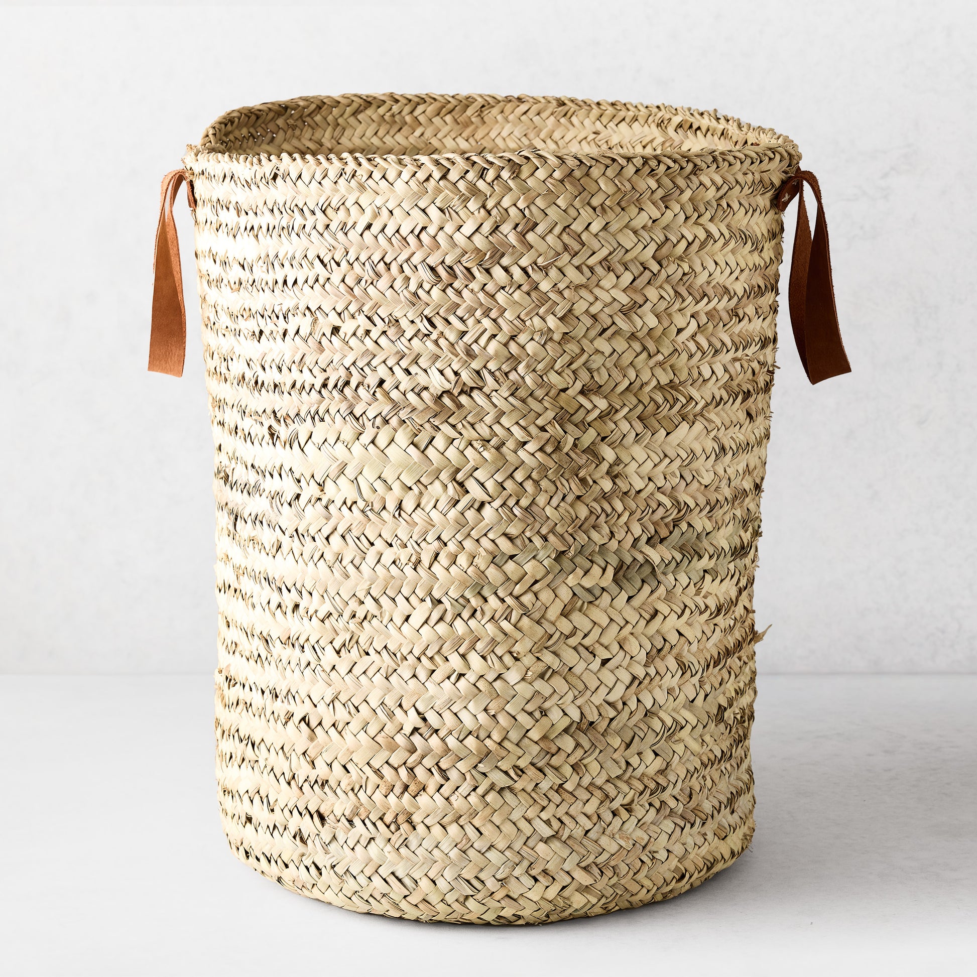 Woven laundry basket made from natural palm grass with brown leather handles.