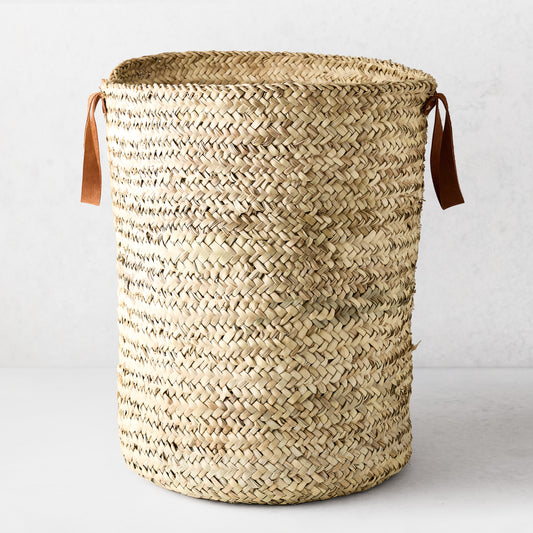 Woven laundry basket made from natural palm grass with brown leather handles.