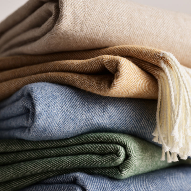 Stack of Italian herringbone throw blankets in blue, green, beige, and gold with fringe.