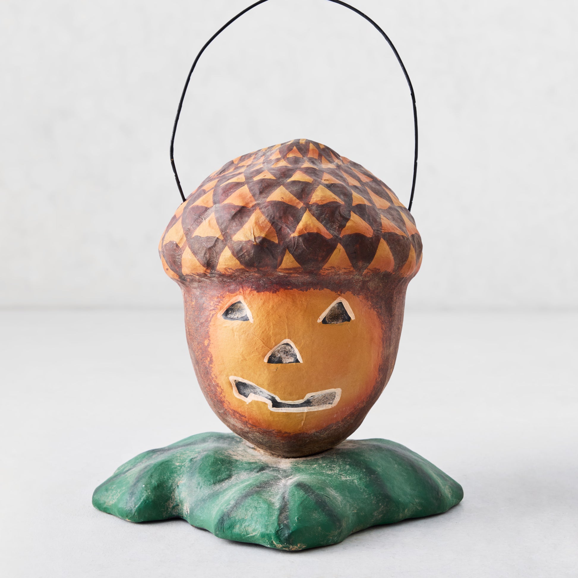 Distressed papier-mâché acorn Halloween candy bucket with a jack-o'-lantern face, green leaf base, and wire handle.