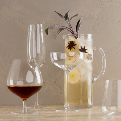 HG Heritage Cognac Glass with amber liquid, alongside other clear drinkware and a pitcher with apple slices.