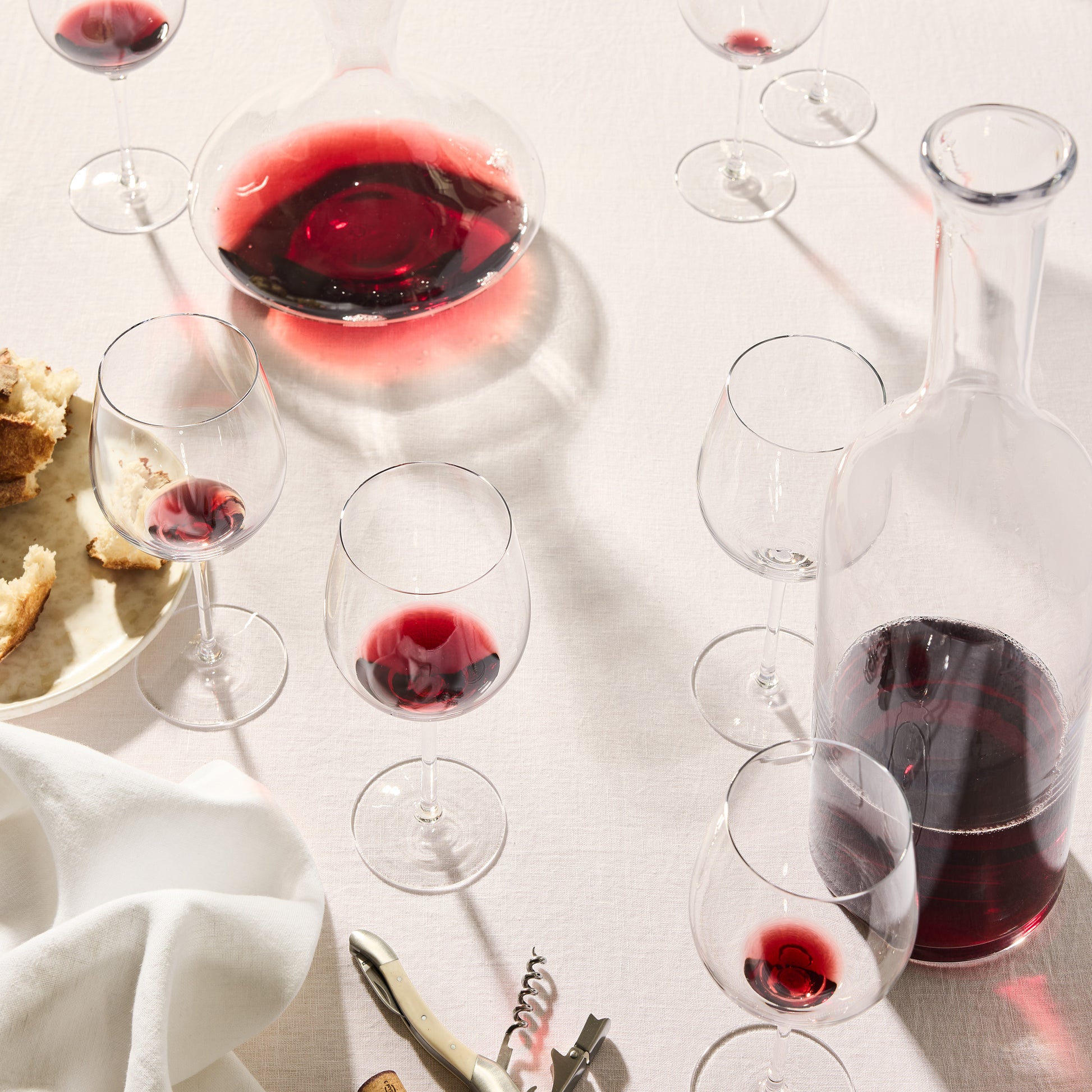 Clear, thin, mouth-blown HG Heritage Wine Glasses and decanters filled with red wine on a white tablecloth.