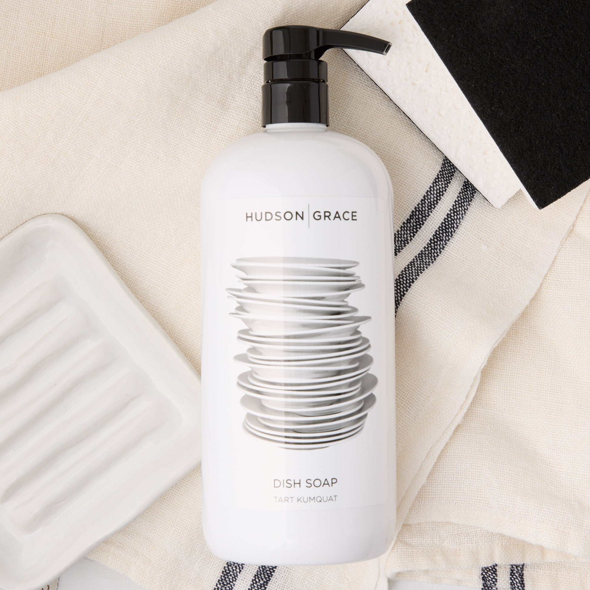 Hudson Grace Tart Kumquat dish soap, white ceramic soap dish, black and white sponges on a linen towel.