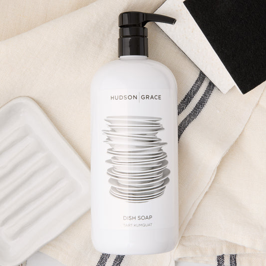 Hudson Grace Tart Kumquat dish soap, white ceramic soap dish, black and white sponges on a linen towel.
