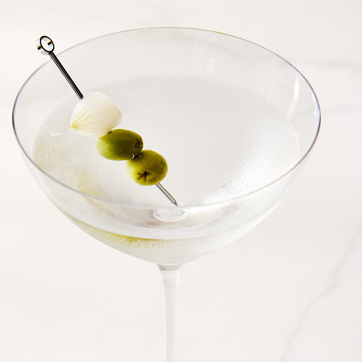 Oversized mouth-blown glass martini glass with a cocktail pick featuring olives and a pearl onion.