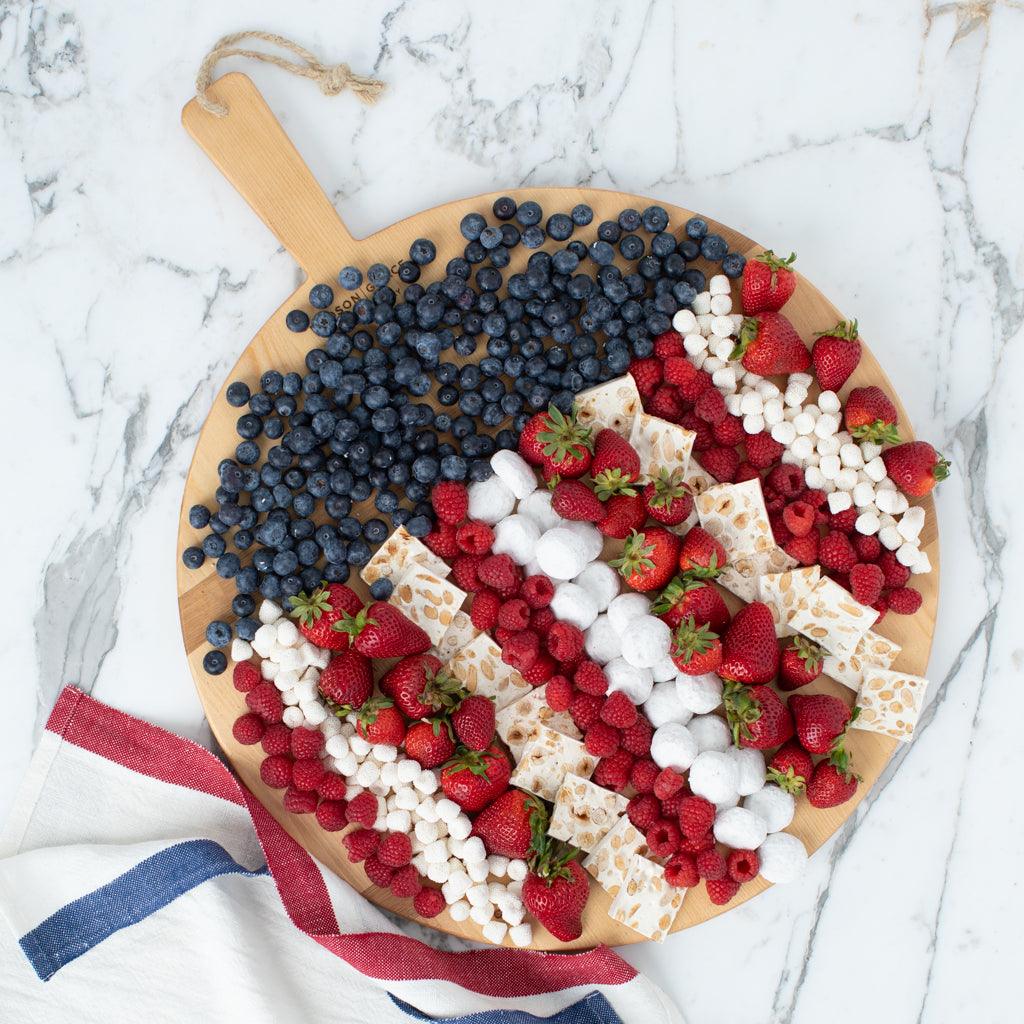 Round reclaimed wood serving board with handle, filled with blueberries, strawberries, raspberries, marshmallows, and nougat.