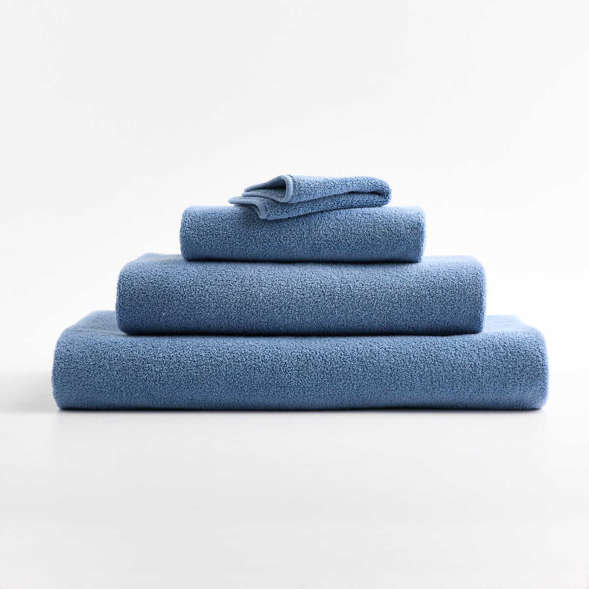 Stack of soft denim blue zero-twist cotton towels in varying sizes with piped edges on a white background.