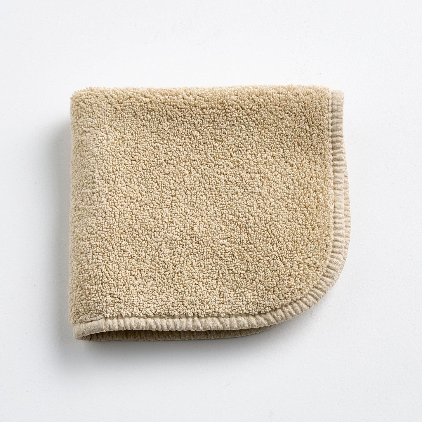 Folded sand-colored Signature washcloth in plush cotton terry with a rounded corner and stitched hem.