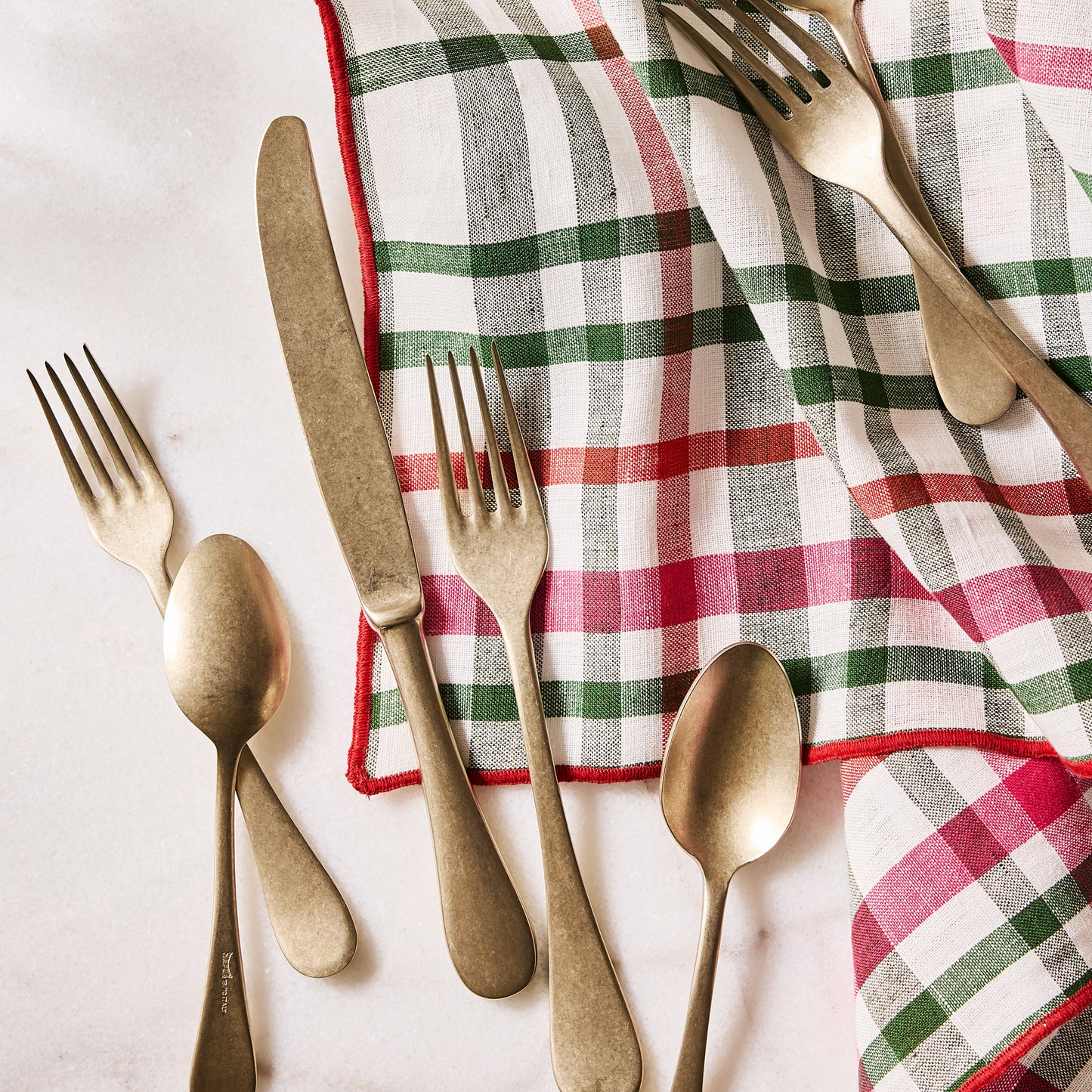 Holiday green and red gingham cotton napkin with embroidered red trim, folded next to gold flatware.