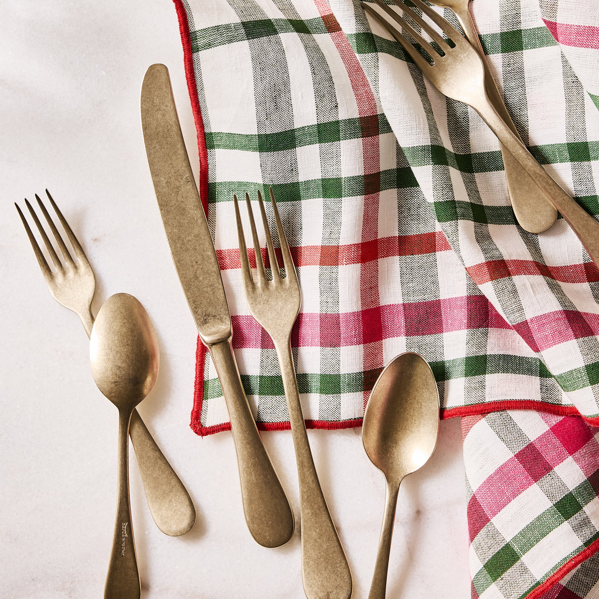 Holiday green and red gingham cotton napkin with embroidered red trim, folded next to gold flatware.