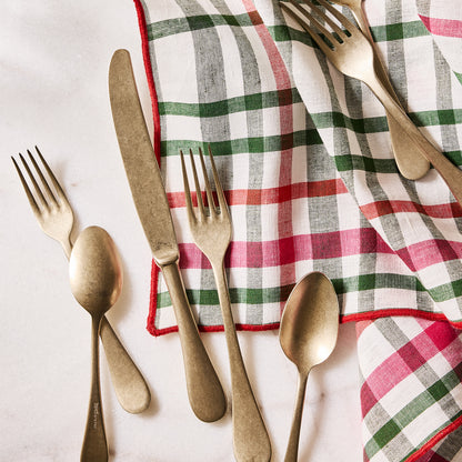Holiday green and red gingham cotton napkin with embroidered red trim, folded next to gold flatware.