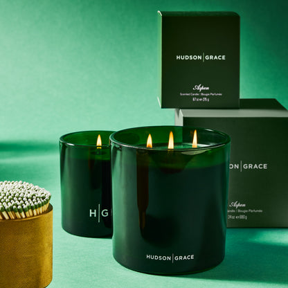 Hudson Grace Aspen Scented 3-Wick Candle in a dark green glass vessel with lit wicks, next to a smaller candle and packaging.
