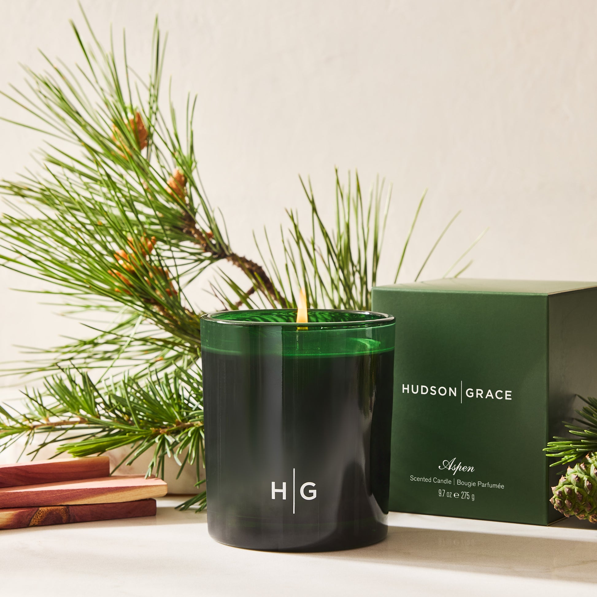 Hudson Grace Aspen Scented Candle in a dark green glass vessel with a burning wick, beside its matching box and pine branches.