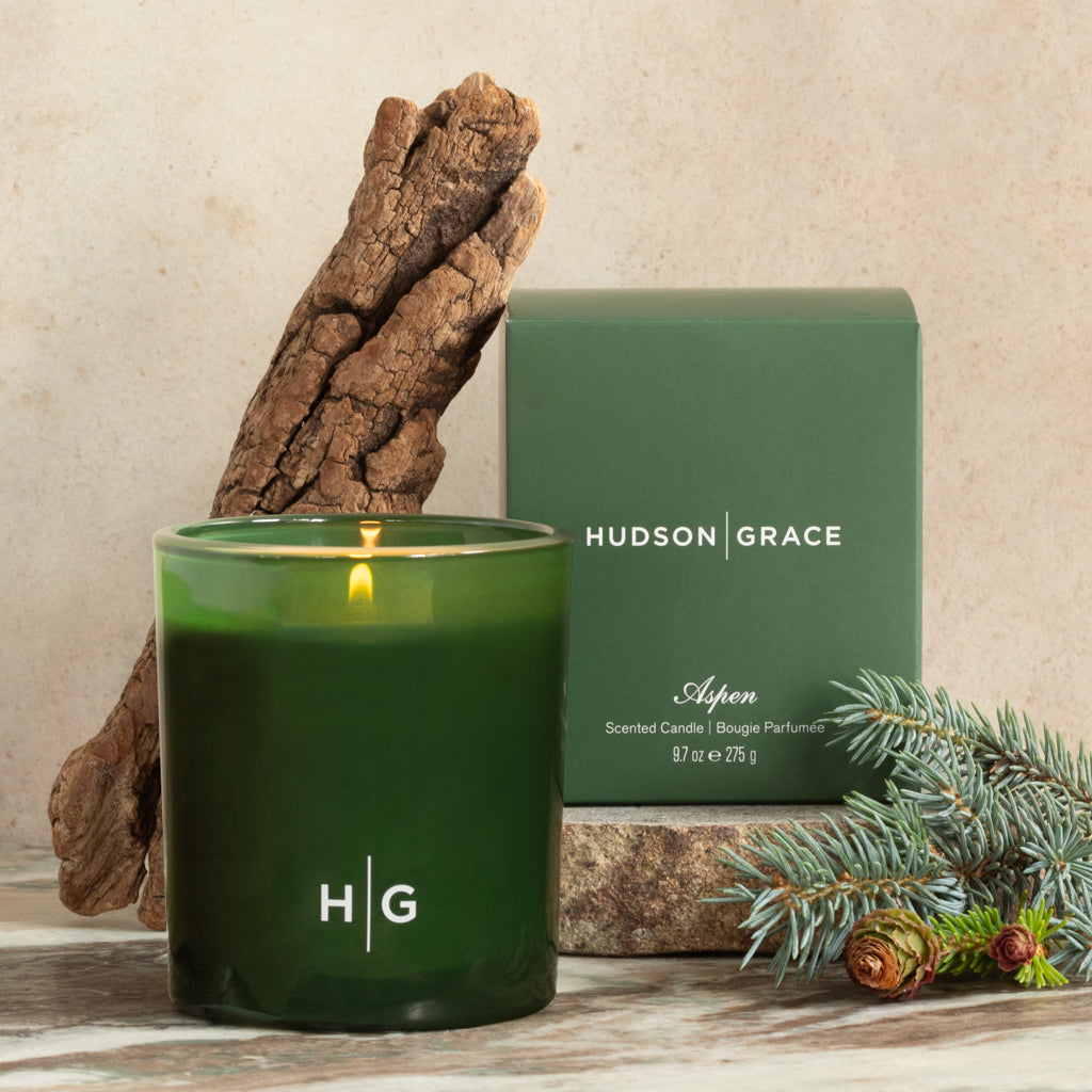 Hudson Grace Aspen green glass scented candle with burning wick, matching box, and fir sprig.