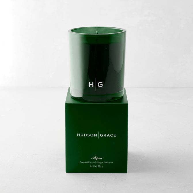 Hudson Grace Aspen Scented Candle in a dark green glass vessel with "HG" logo, atop its matching green box.