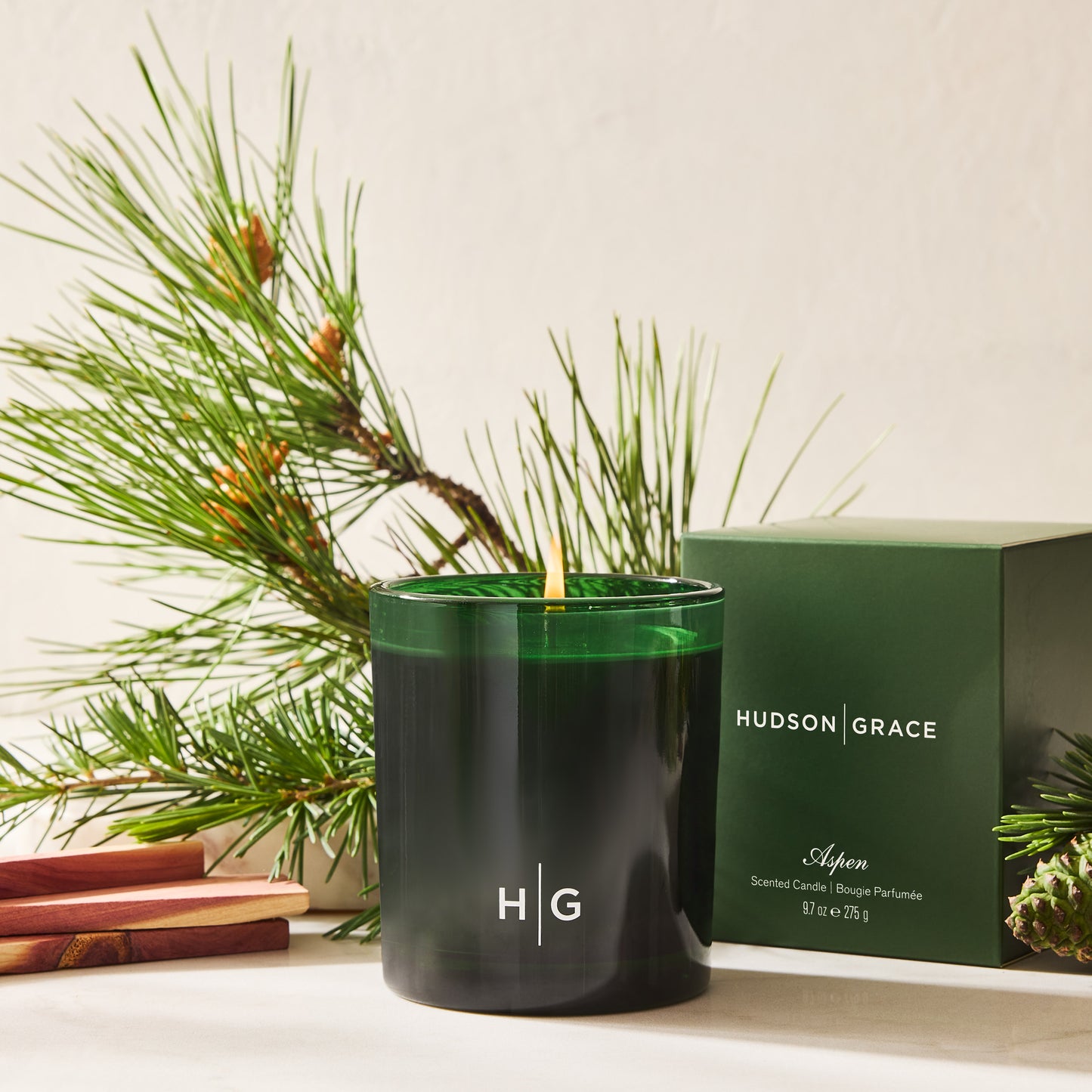 Hudson Grace Aspen scented candle in a dark green glass votive with a lit wick, next to its green box.