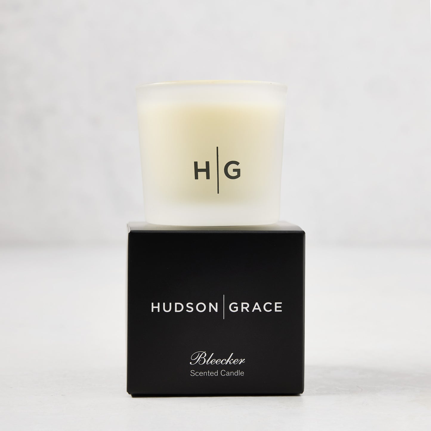 Hudson Grace Bleecker Scented Votive in a frosted glass vessel with HG logo, atop a black box.