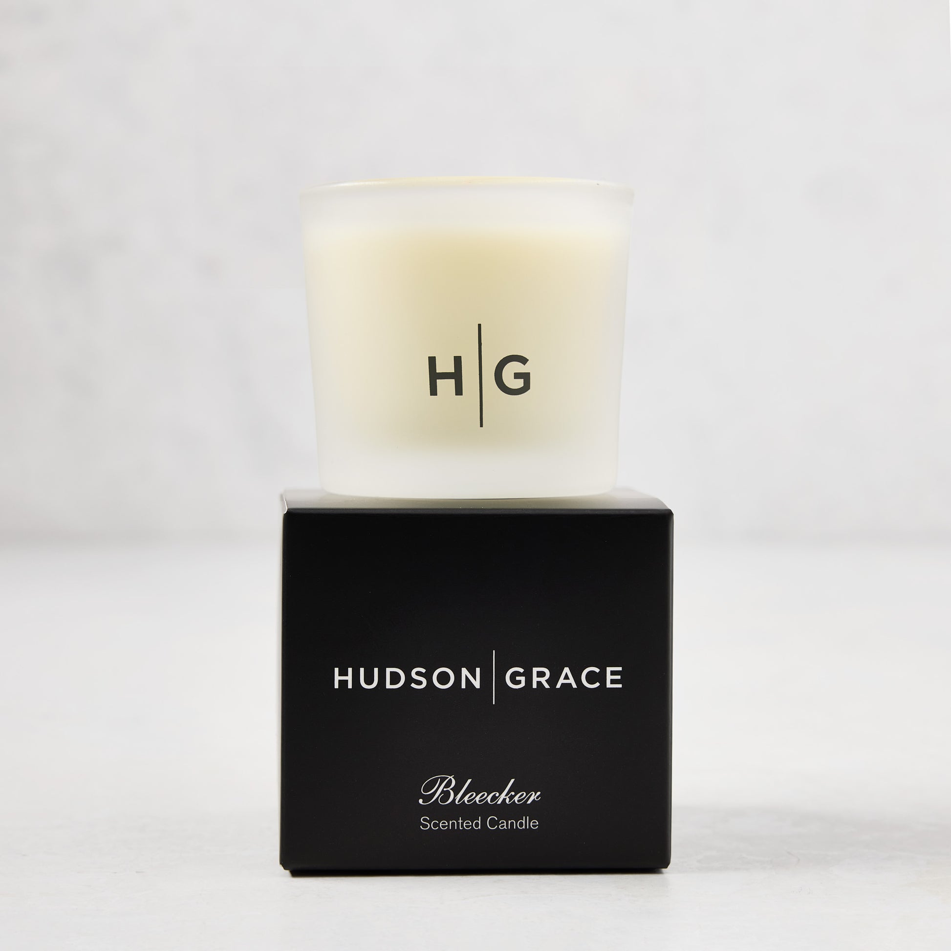 Hudson Grace Bleecker Scented Votive in a frosted glass vessel with HG logo, atop a black box.