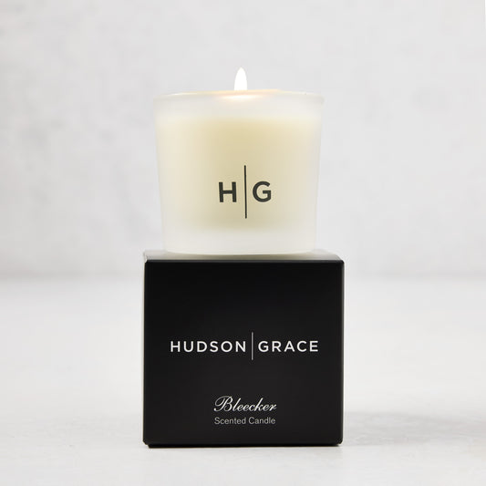 Hudson Grace Bleecker Scented Votive, a lit candle in a frosted glass vessel with "HG" logo, on a black box.