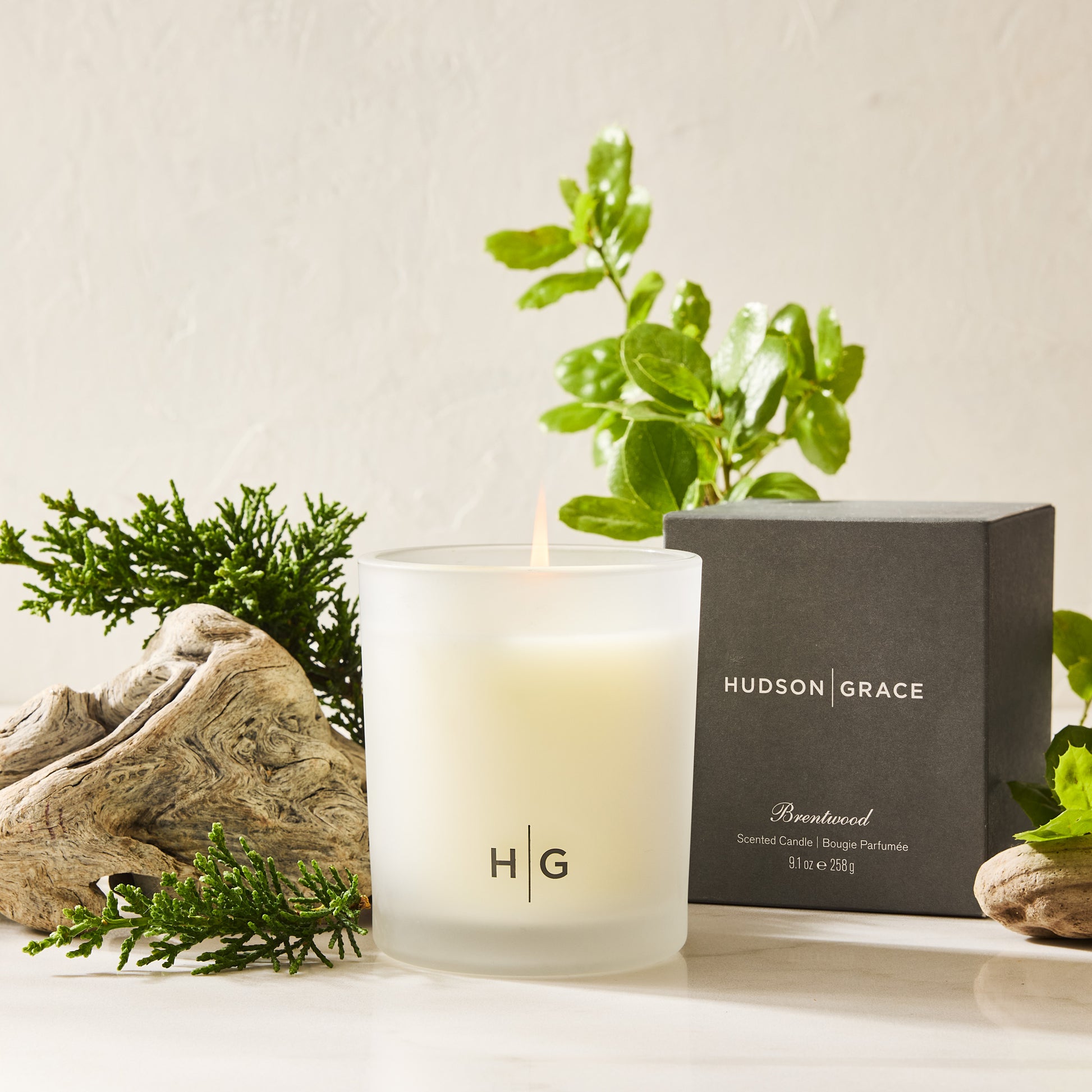 Hudson Grace Brentwood scented candle in a frosted glass vessel with a lit wick, next to its black gift box.