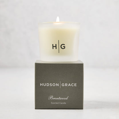 Hudson Grace Brentwood Votive Scented Candle in a frosted glass vessel, lit, on a dark gray box.