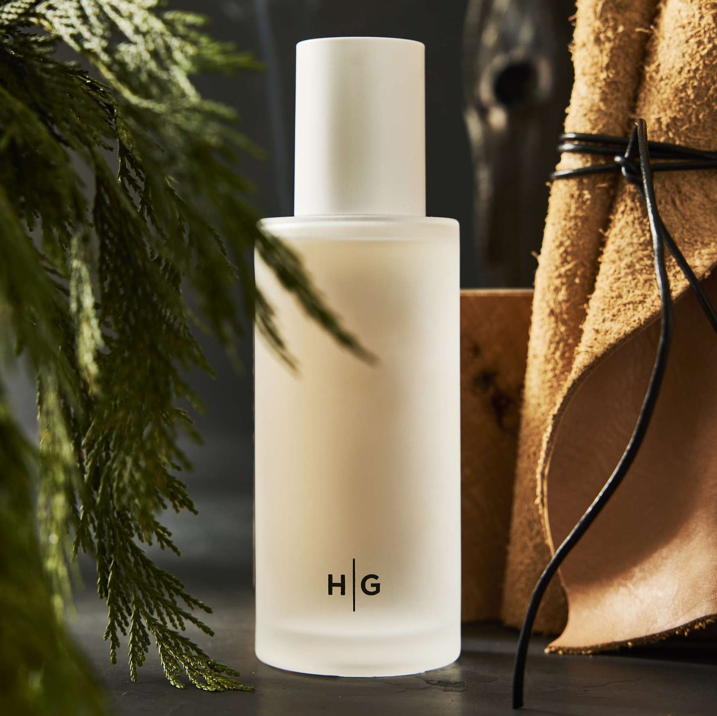Frosted glass Hudson Grace Aspen candle with green fir branches, brown leather, and dark background.