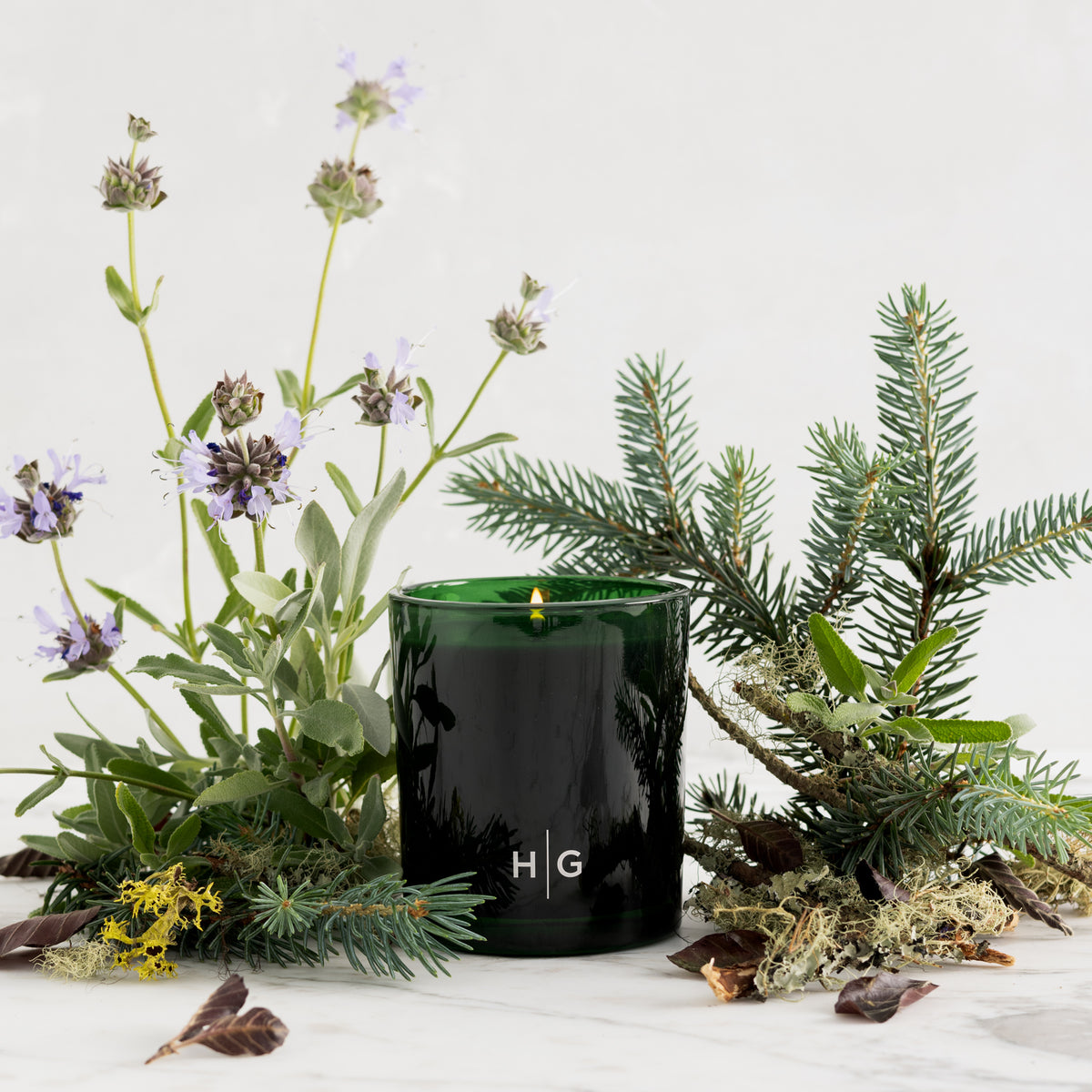 Hudson Grace Aspen candle in a dark green glass jar, surrounded by sage flowers, pine branches, and lichen on marble.