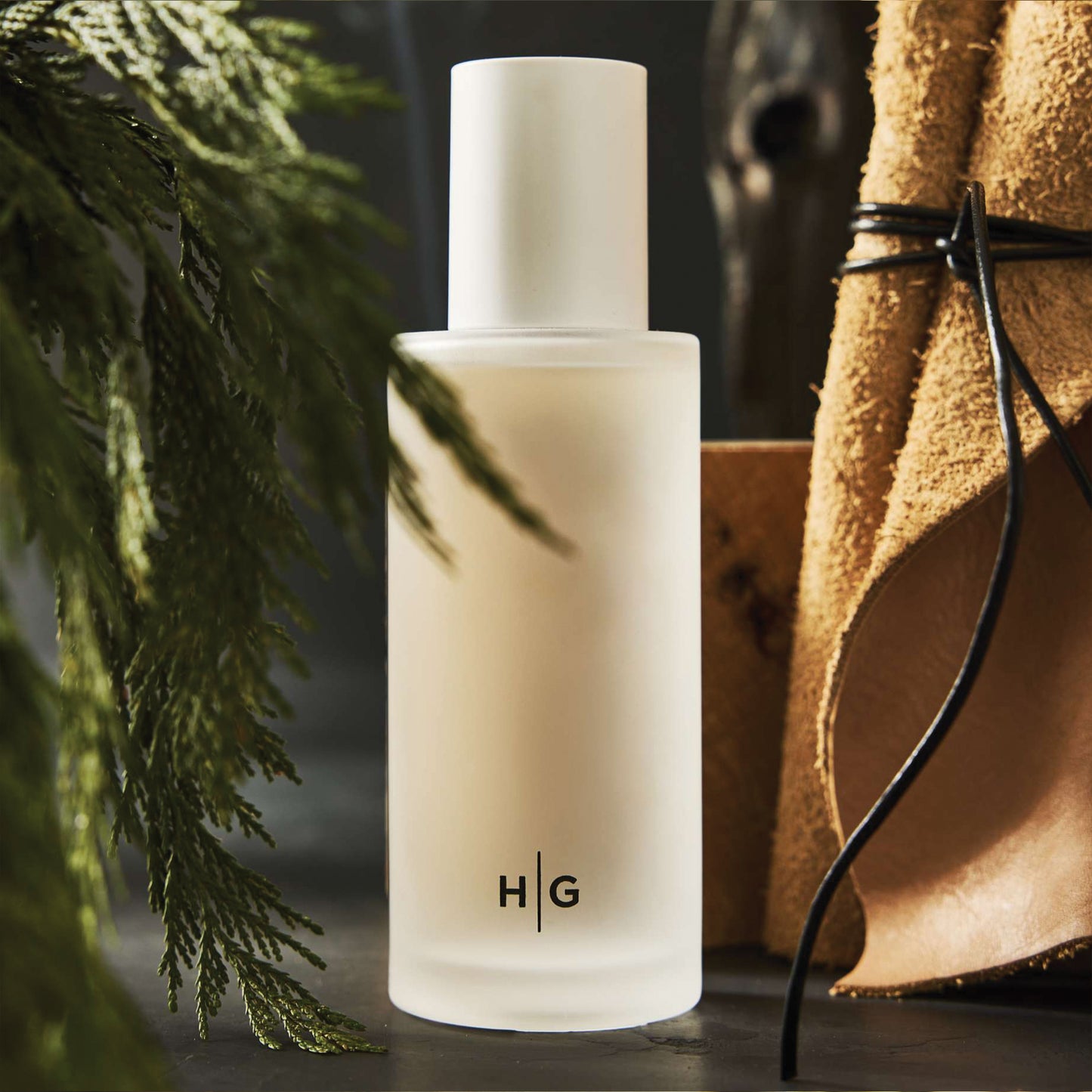 Hudson Grace Bleecker fragrance bottle with HG logo, surrounded by green foliage and brown leather.