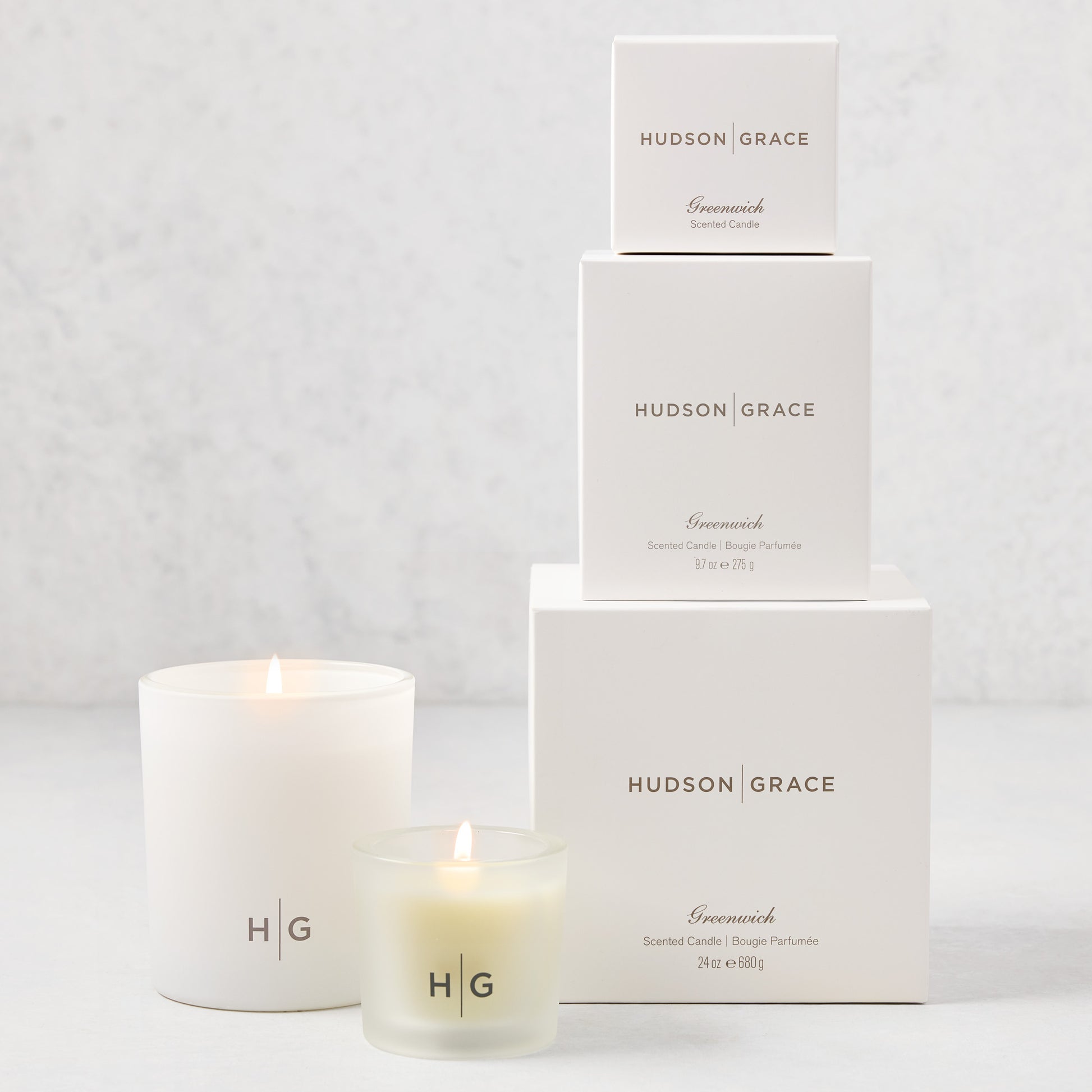 Hudson Grace Greenwich Candle Gift Set: 3 white boxes stacked, 2 lit candles (votive, classic) with HG logo.