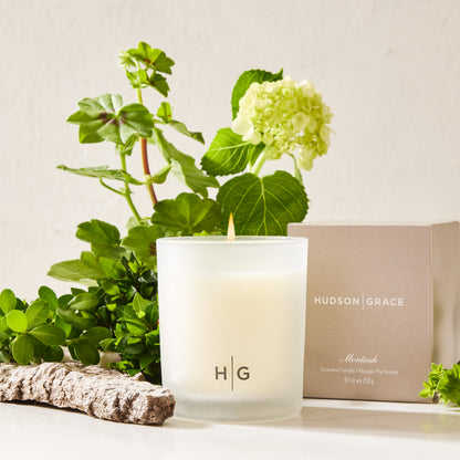 Hudson Grace Montauk scented candle in a frosted glass jar with a lit wick, next to its gift box and green plants.