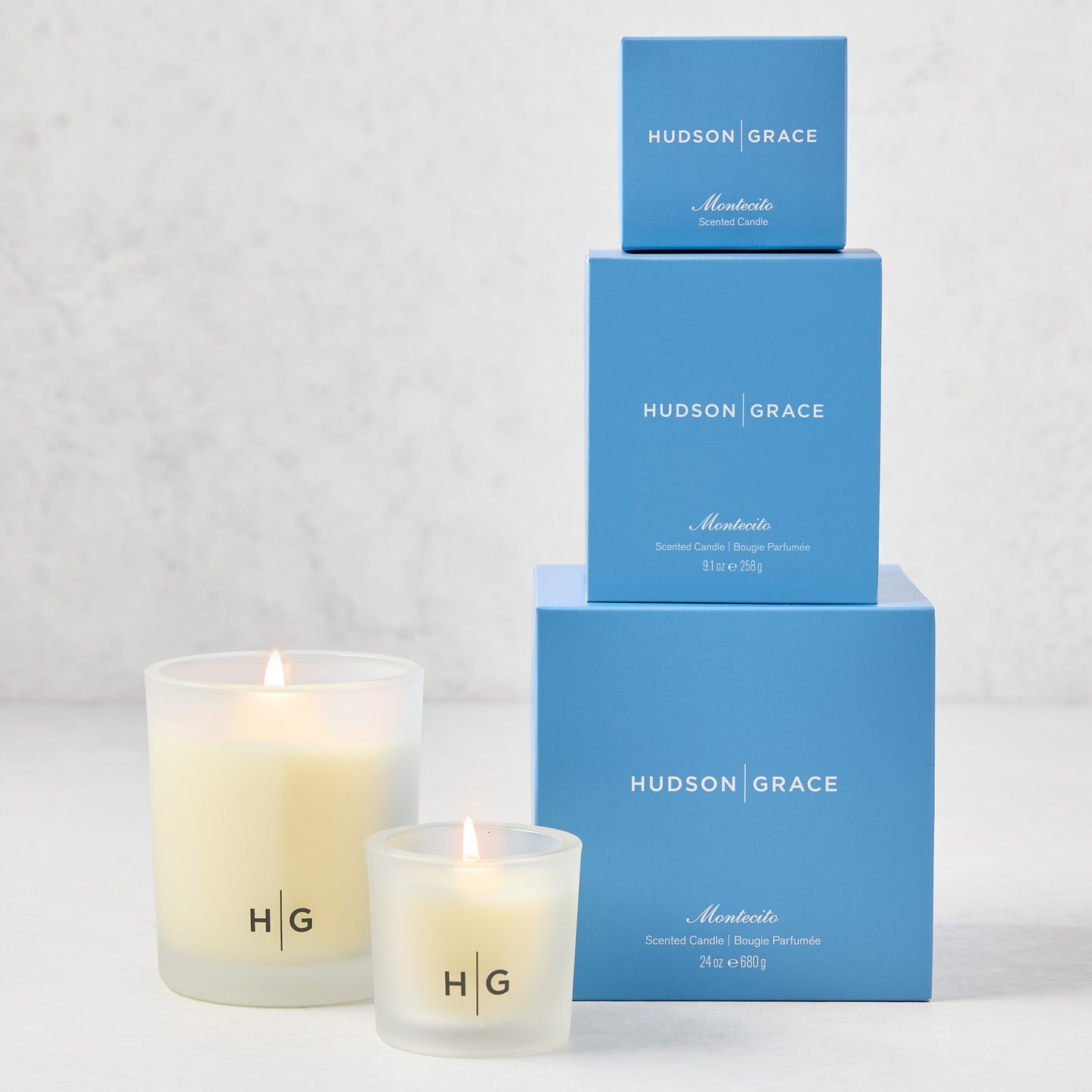 Hudson Grace Montecito Candle Gift Set: 3 blue boxes stacked, 2 lit frosted glass candles with "HG" logo.