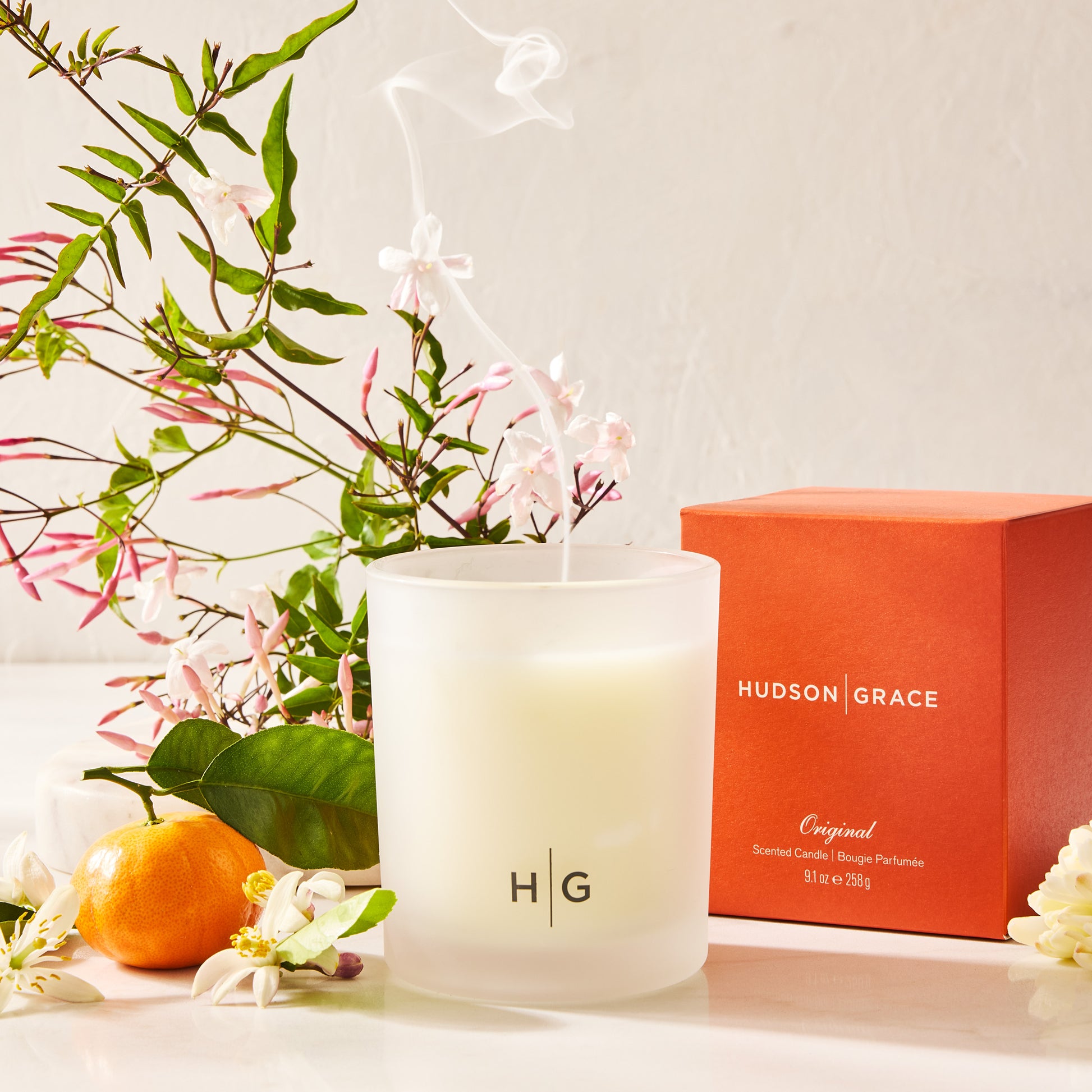Hudson Grace Original scented candle in a frosted glass jar with an orange box and jasmine flowers.
