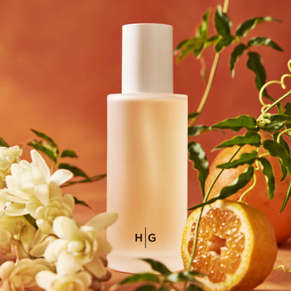 Hudson Grace Original Candle, frosted glass bottle with white cap, surrounded by white flowers and sliced oranges.