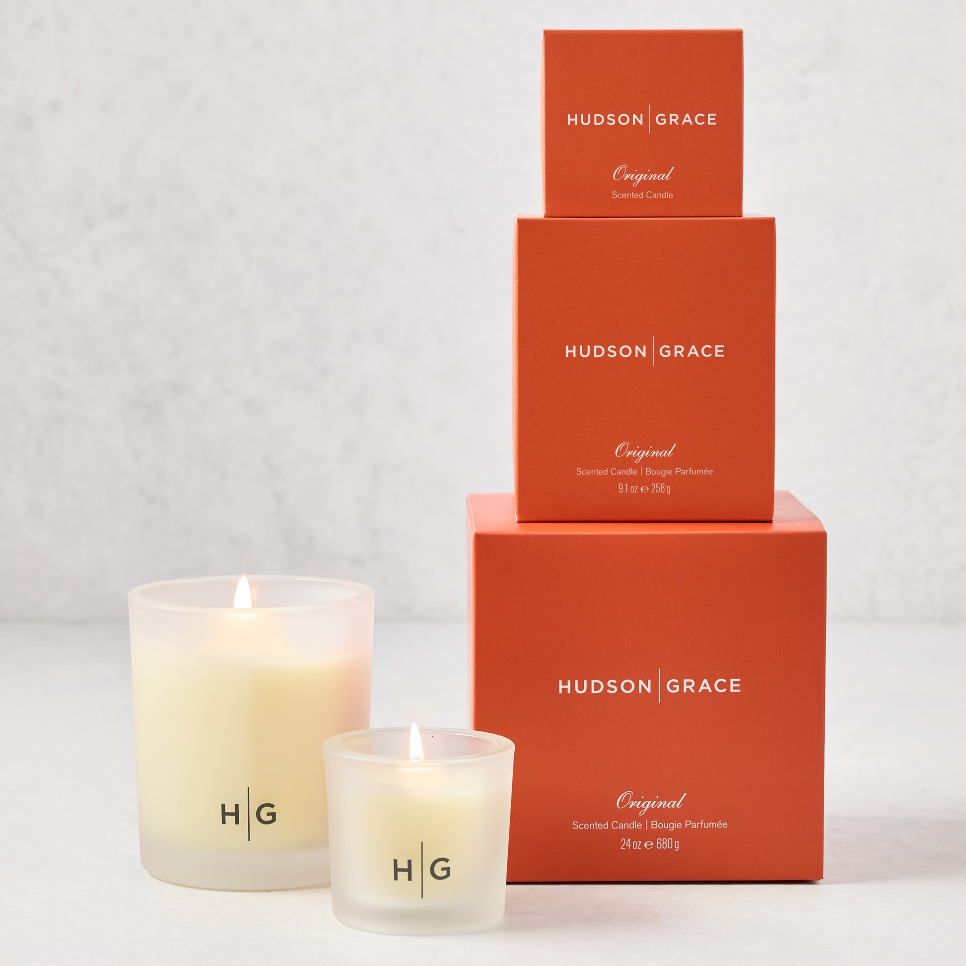 Hudson Grace Original Candle Gift Set: 3 lit candles in frosted glass jars, 3 stacked orange boxes.