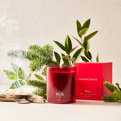Red Hudson Grace Winter candle in a glass jar, lit with smoke, next to its red box, pine, and greenery.