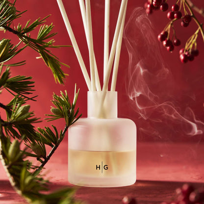 Hudson Grace Winter reed diffuser with white reeds, HG logo, pine, and red berries on a red background.