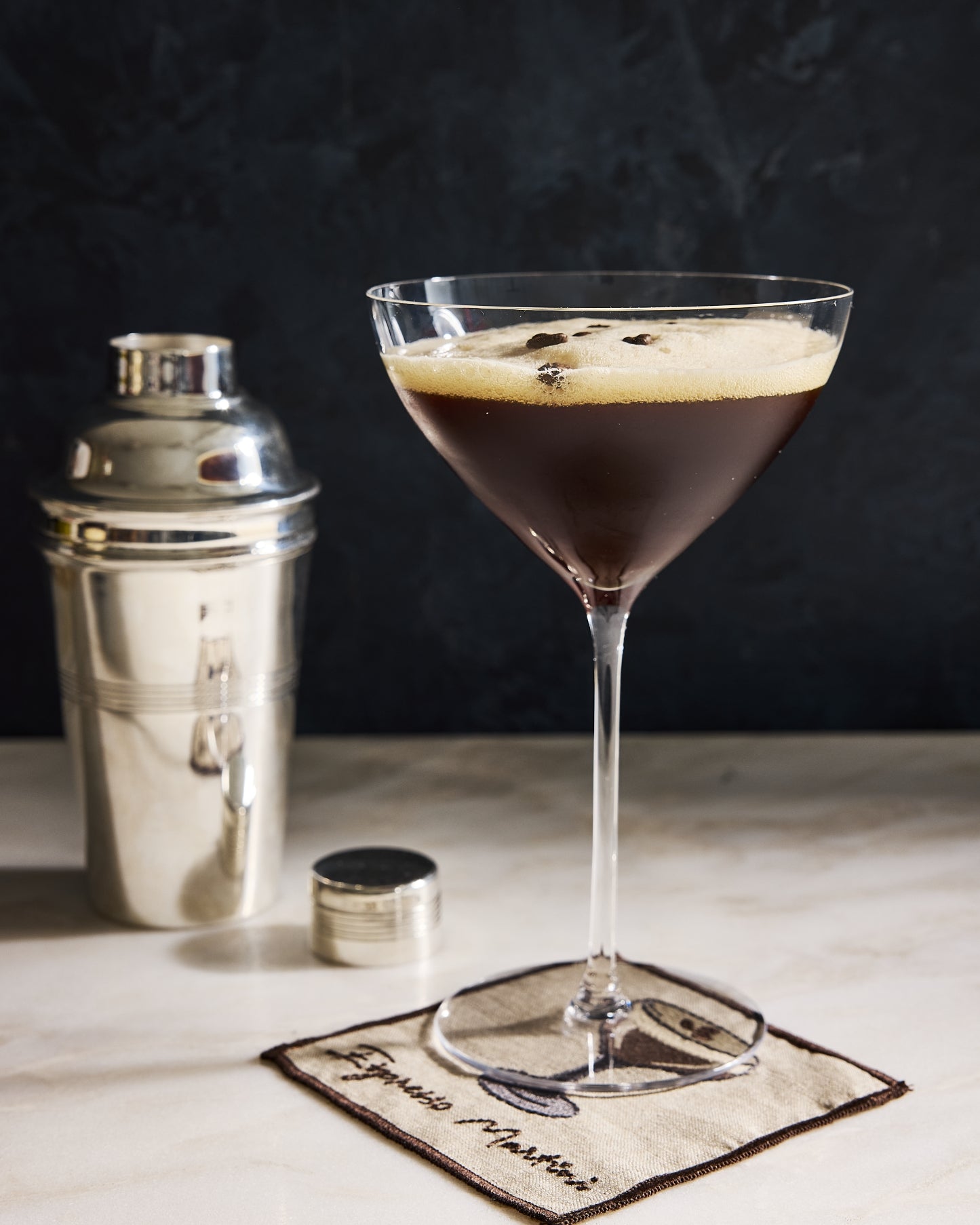 A martini glass filled with a dark espresso-based cocktail and a foamy top, set on a napkin labeled Espresso Martini, with a metal cocktail shaker and small tin on a light marble surface against a dark textured background.