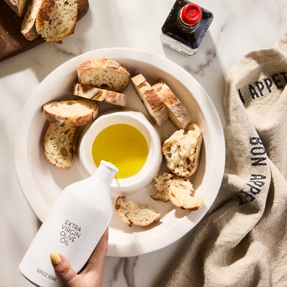 White ceramic Hudson Grace Extra Virgin Olive Oil bottle pouring into a dipping dish with bread slices.