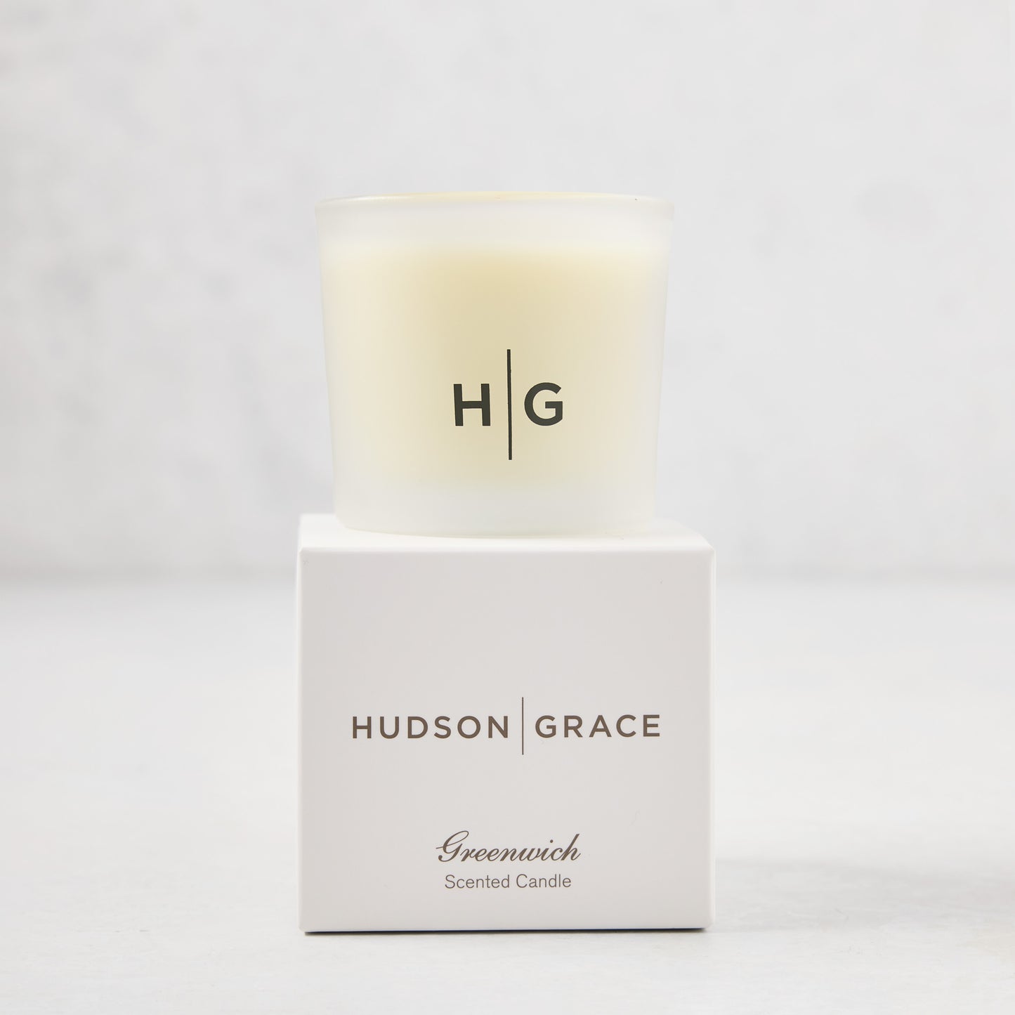 Hudson Grace Greenwich Scented Votive, a small candle in a frosted glass vessel with "HG" logo, on its white box.