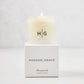 Hudson Grace Greenwich Scented Votive, a lit candle in a frosted glass vessel with "HG" logo, on its white box.